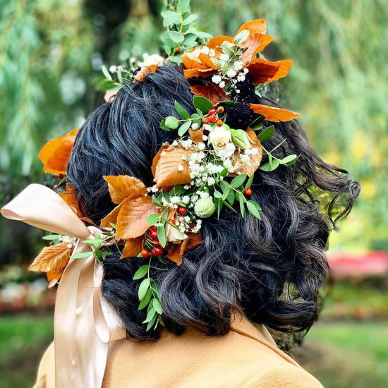 The image showcases the back of a person’s head adorned with a beautiful arrangement of flowers and leaves, creating a natural and elegant look. The individual has dark, curly hair that provides a contrasting backdrop to the vibrant colors of the foliage and blossoms intertwined within it. The setting appears to be outdoors, surrounded by greenery. Order online for wedding & event flowers from the best florist in Toronto near you.