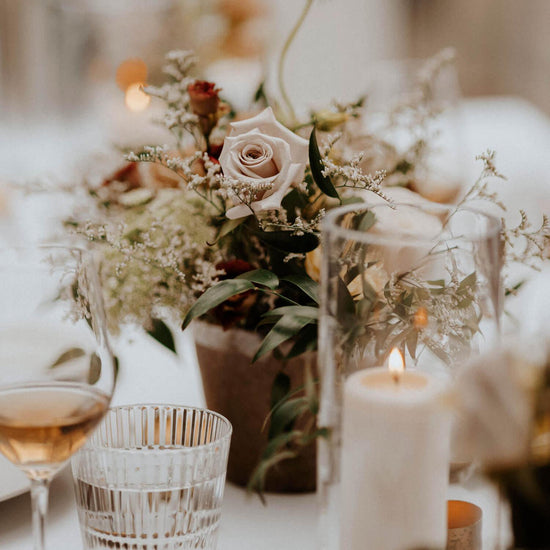 A close-up of a table setting featuring a floral centerpiece, lit candles, and glassware. Order online for wedding & event flowers from the best florist in Toronto near you.
