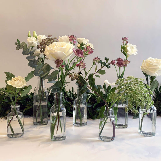 A serene arrangement of white roses, assorted flowers, and greenery in glass vases, accentuated by the soft glow of candles in crystal holders against a light backdrop. Order online for same-day flower delivery from the best wedding florist in Toronto near you.
