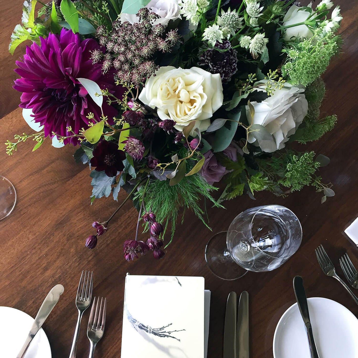 A beautifully arranged bouquet of white, pink, and deep purple flowers accompanied by lush greenery sits in a grey vase on a table set with clear glassware, against the backdrop of an elegant room where guests gather around.  Order online for wedding & event flowers from the best florist in Toronto near you.