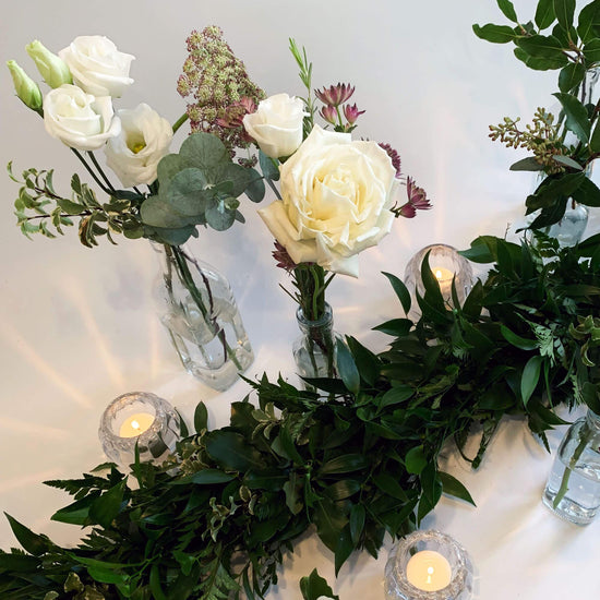 A serene arrangement of white roses, assorted flowers, and greenery in glass vases, accentuated by the soft glow of candles in crystal holders against a light backdrop. Order online for same-day flower delivery from the best wedding florist in Toronto near you.