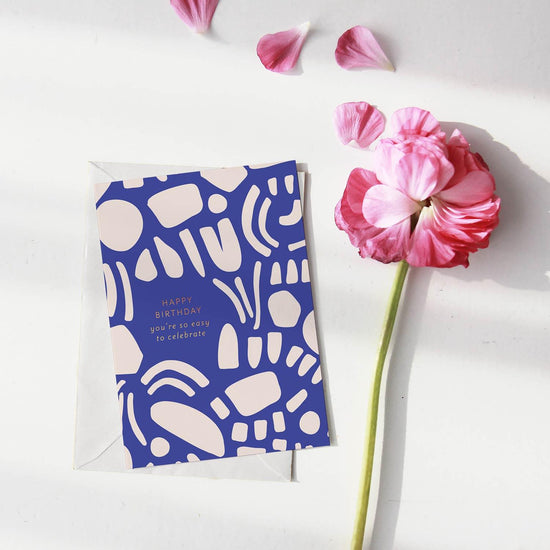 A birthday greeting card with a blue background featuring abstract white shapes and a birthday message in white text: ‘HAPPY BIRTHDAY you’re so easy to celebrate’.Order online for event items from the best florist in Toronto near you.