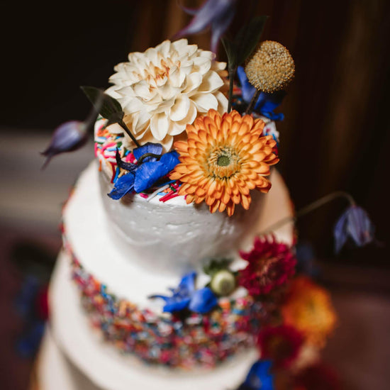 Top tier of a white cake beautifully adorned with a vibrant assortment of flowers including a large cream-colored flower, an orange gerbera, and blue petals, surrounded by other colorful floral decorations. Order online for same-day flower delivery from the best florist in Toronto near you.