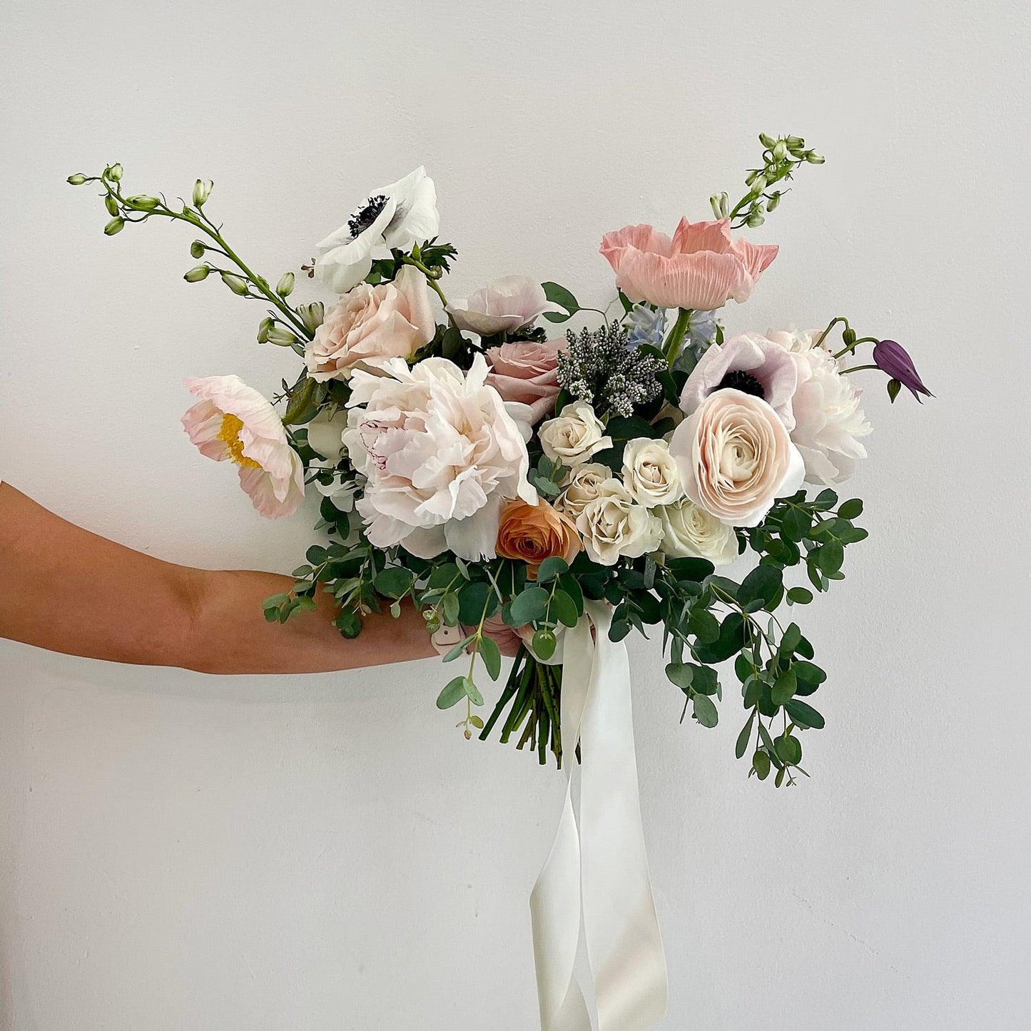 Beautiful bridal bouquet of assorted flowers including roses and poppies, adorned with a white ribbon, against a plain white background. Our bouquets marry creative vision and nature. We work with luxurious flower varieties including garden roses, dahlias, and peonies in season. They emphasize blooms over greenery, are hand-tied & finished with a ribbon of your choice, and come in a vase. From the best wedding florist in Toronto Quince Flowers.