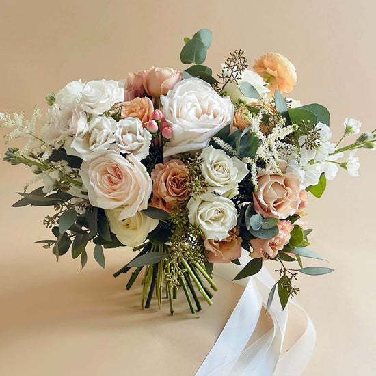 Beautiful bridal bouquet of assorted flowers including roses and poppies, adorned with a white ribbon, against a plain white background.  Order online for wedding & event flowers from the best florist in Toronto near you.