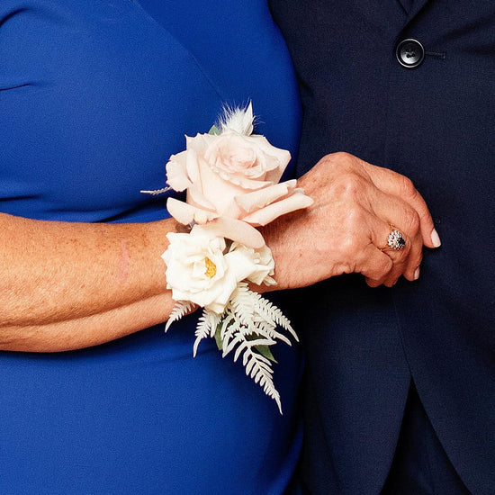 A close-up image of two people, likely at a formal event, with their arms interlocked. One person is wearing a corsage made of pale roses and ferns. Order online for wedding & event flowers from the best florist in Toronto near you.