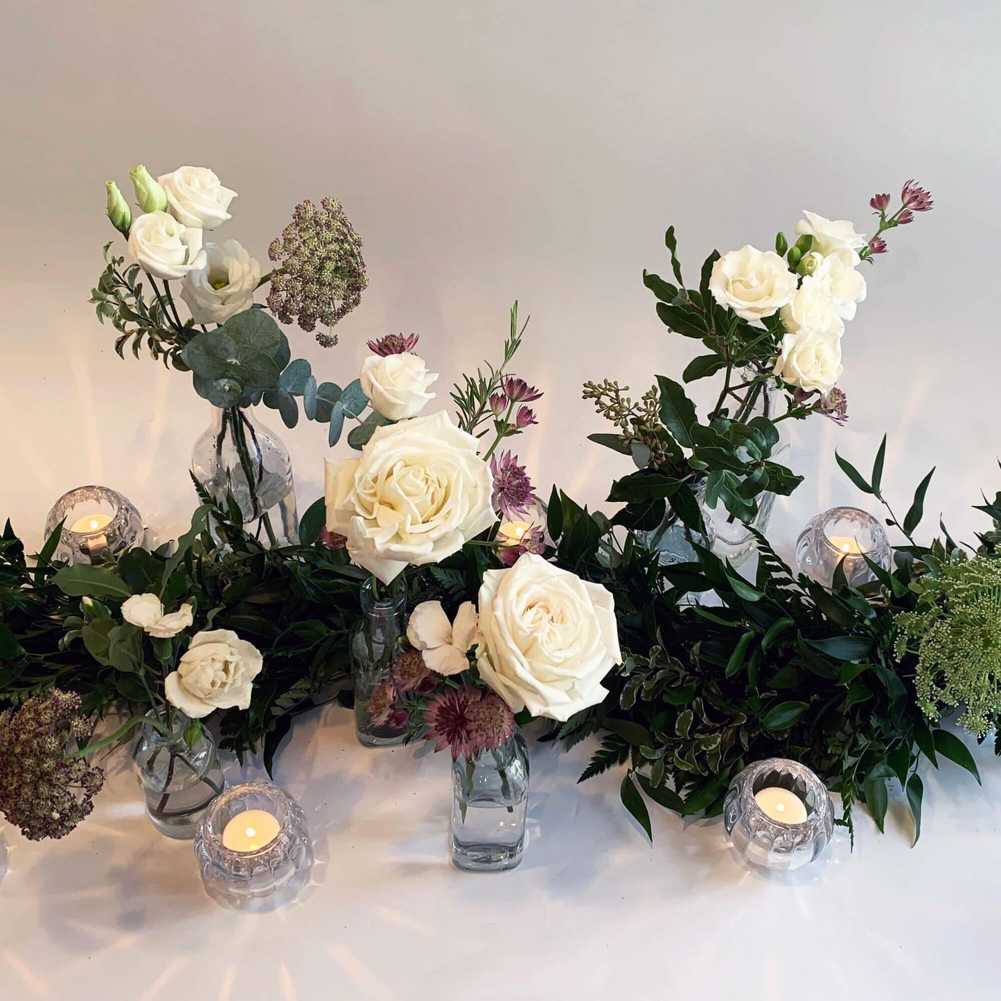 Load image into Gallery viewer, A serene arrangement of white roses, assorted flowers, and greenery in glass vases, accentuated by the soft glow of candles in crystal holders against a light backdrop. Order online for same-day flower delivery from the best wedding florist in Toronto near you.
