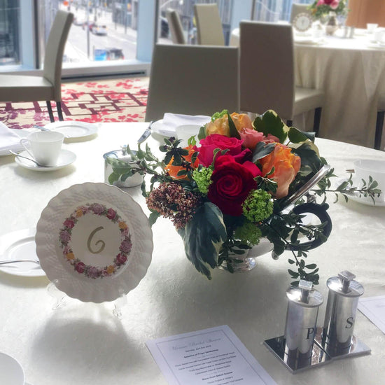 Load image into Gallery viewer, A well-lit dining table set up with a vibrant bouquet of flowers including red roses and orange lilies in a glass vase, a round card with the number 6 surrounded by floral designs, white porcelain teacups and saucers, and salt and pepper shakers. A printed menu is also visible on the table. The background reveals more tables with similar settings and a large window allowing natural light to illuminate the room.  Order online for wedding &amp;amp; event flowers from the best florist in Toronto near you.
