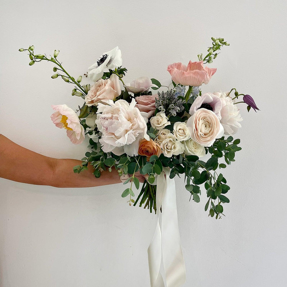 
                      
                        Beautiful bridal bouquet of assorted flowers including roses and poppies, adorned with a white ribbon, against a plain white background. Our bouquets marry creative vision and nature. We work with luxurious flower varieties including garden roses, dahlias, and peonies in season. They emphasize blooms over greenery, are hand-tied & finished with a ribbon of your choice, and come in a vase. From the best wedding florist in Toronto Quince Flowers.
                      
                    