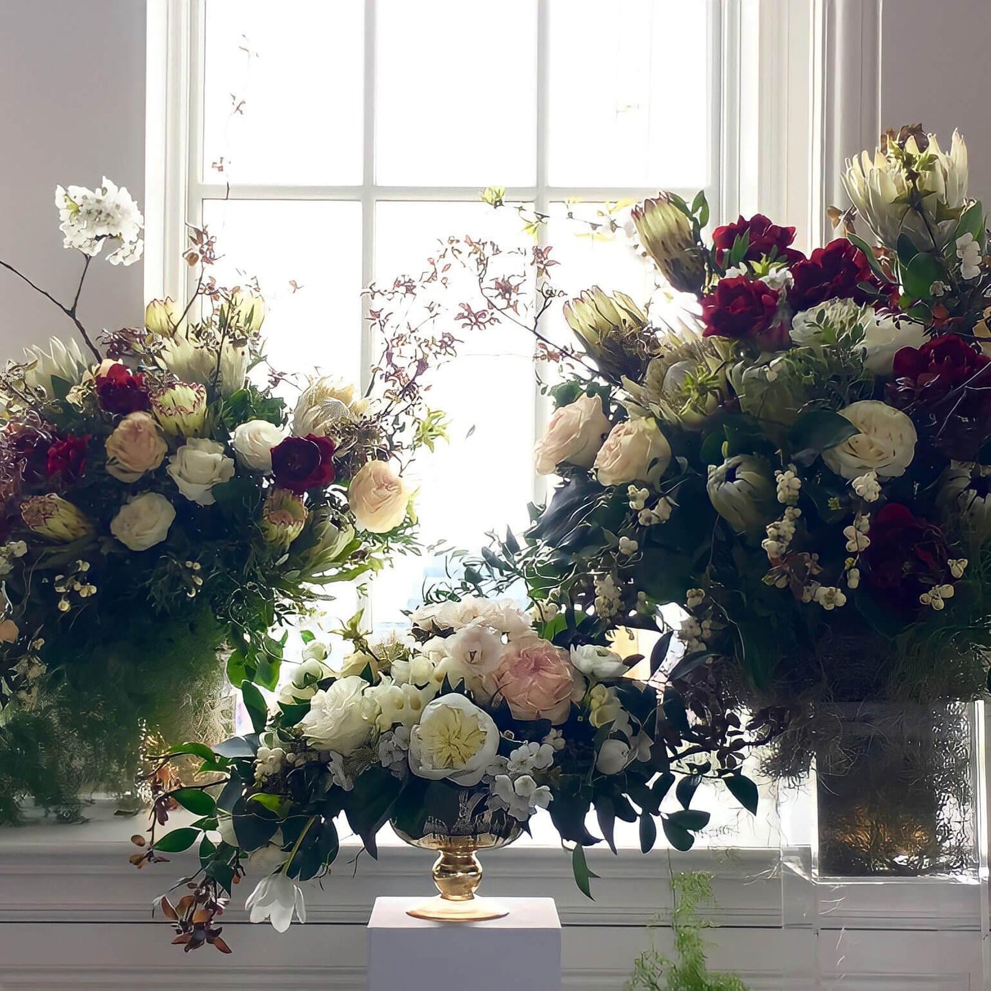 A beautiful arrangement of various flowers in a golden vase, placed on a white pedestal against a window, creating a serene and elegant atmosphere.  Order online for sympathy & event flowers from the best florist in Toronto near you.