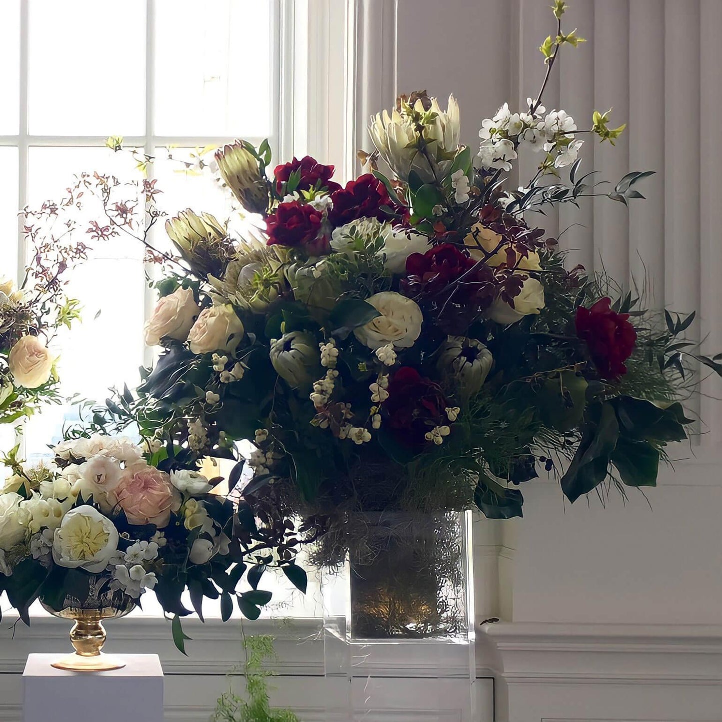 A beautiful arrangement of various flowers in a golden vase, placed on a white pedestal against a window, creating a serene and elegant atmosphere. Order online for sympathy & event flowers from the best florist in Toronto near you.