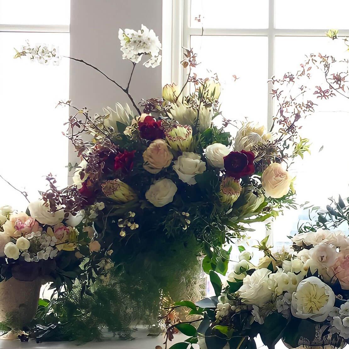 A beautiful arrangement of various flowers in a golden vase, placed on a white pedestal against a window, creating a serene and elegant atmosphere. Order online for sympathy & event flowers from the best florist in Toronto near you.
