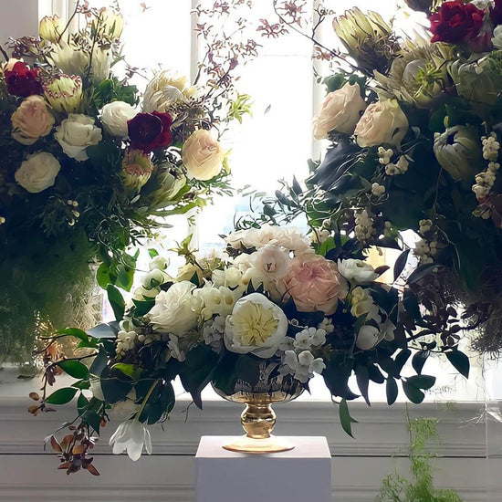 A beautiful arrangement of various flowers in a golden vase, placed on a white pedestal against a window, creating a serene and elegant atmosphere. Order online for sympathy & event flowers from the best florist in Toronto near you.
